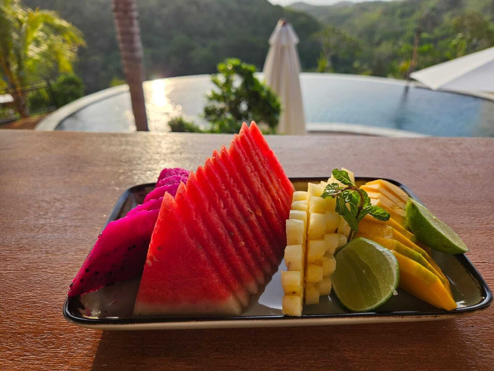 A fruit platter on a table with a pool and mountains in the background.