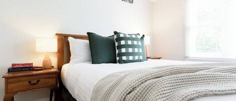 A neatly made bed with a white comforter and a beige knitted throw. There are two pillows, one green and one with a checkered pattern. A bedside table holds a lamp and a stack of books. The room has a window with blinds, and artwork is displayed on the wall.