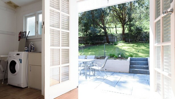 A laundry area with a washing machine, a small cabinet, and a window. Outside, there is a patio with a table and chairs, a grassy yard, and a wooden fence. - The Basin Cottage - Closest Home to The Gorge (West Launceston)