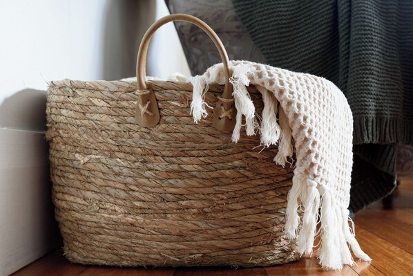A woven basket with a handle and a white fringe, placed on a wooden floor next to a wall.