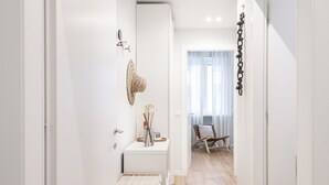 Un pasillo estrecho con una puerta blanca, una mesita pequeña con un florero y una vela, y un cajón de madera.