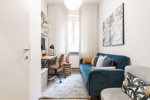 Un salon chaleureux avec un canapé bleu, un fauteuil gris et un tapis blanc. On y trouve un bureau avec une chaise et une lampe. Une fenêtre avec des rideaux est visible au fond.