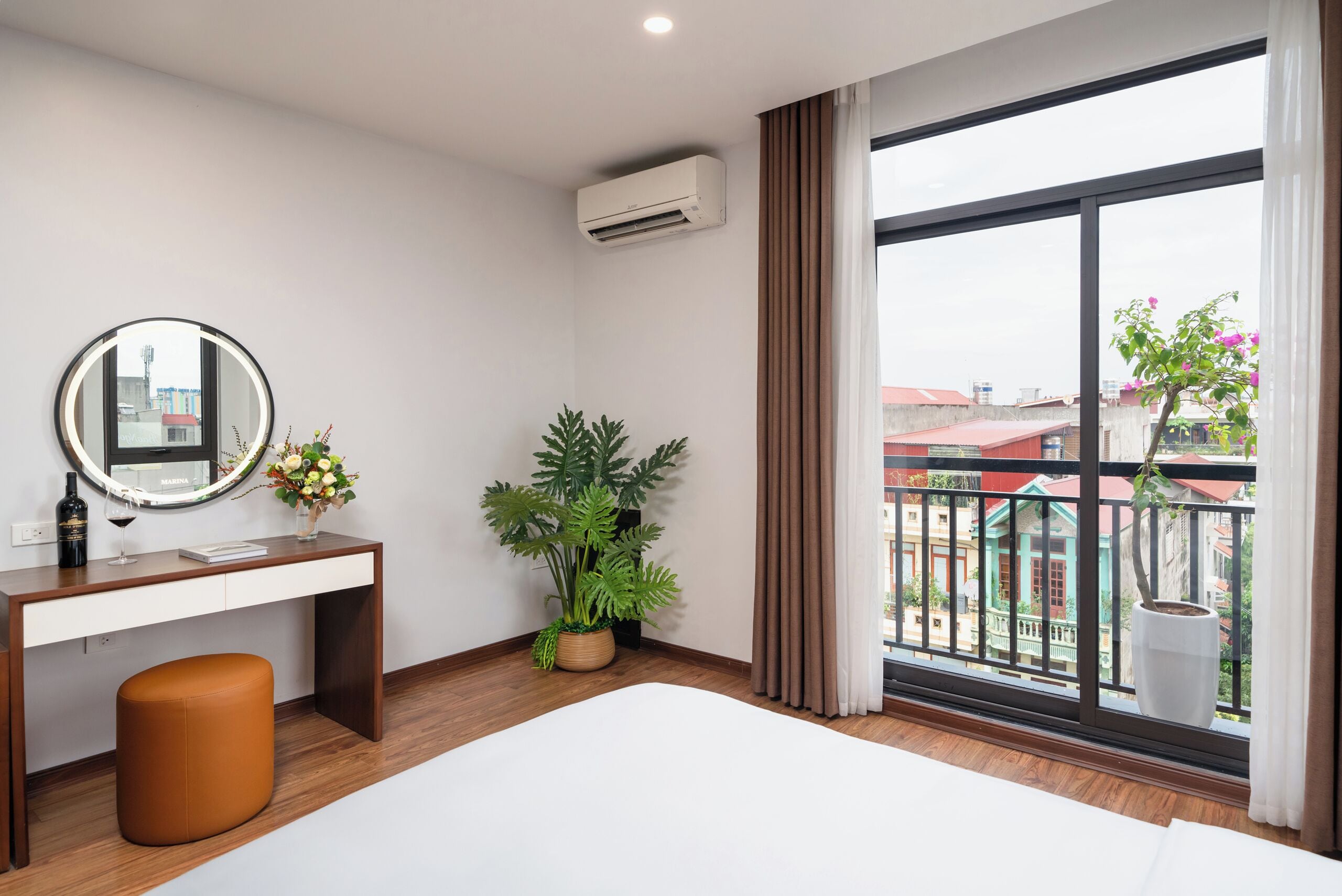 A hotel room with a bed, a desk with a mirror, a potted plant, and a view of buildings through large windows.