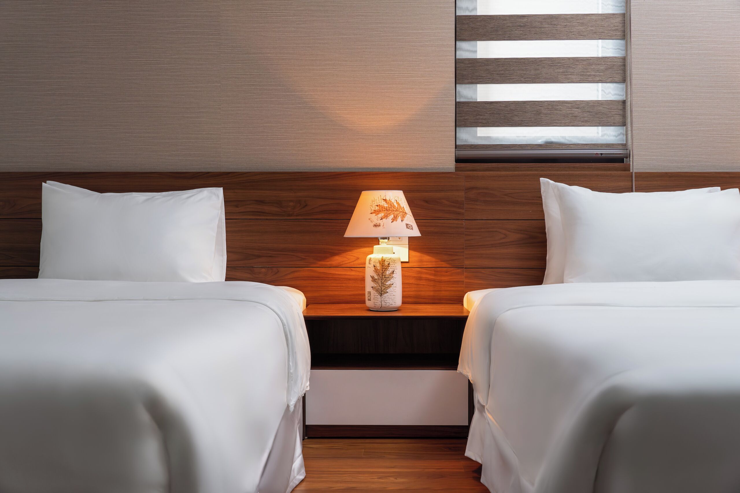 Habitación de hotel con dos camas, una mesita de noche con lámpara y una ventana con persianas.