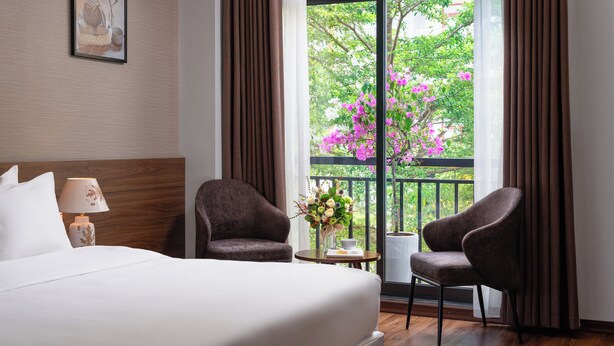 A hotel room with a large bed, two armchairs, a small table with a vase of flowers, and a window with a view of trees and pink flowers. - Marina Hotel (Bac Giang)
