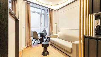 A modern hotel room with a sofa, a small round table, a desk, and a dining area with chairs.