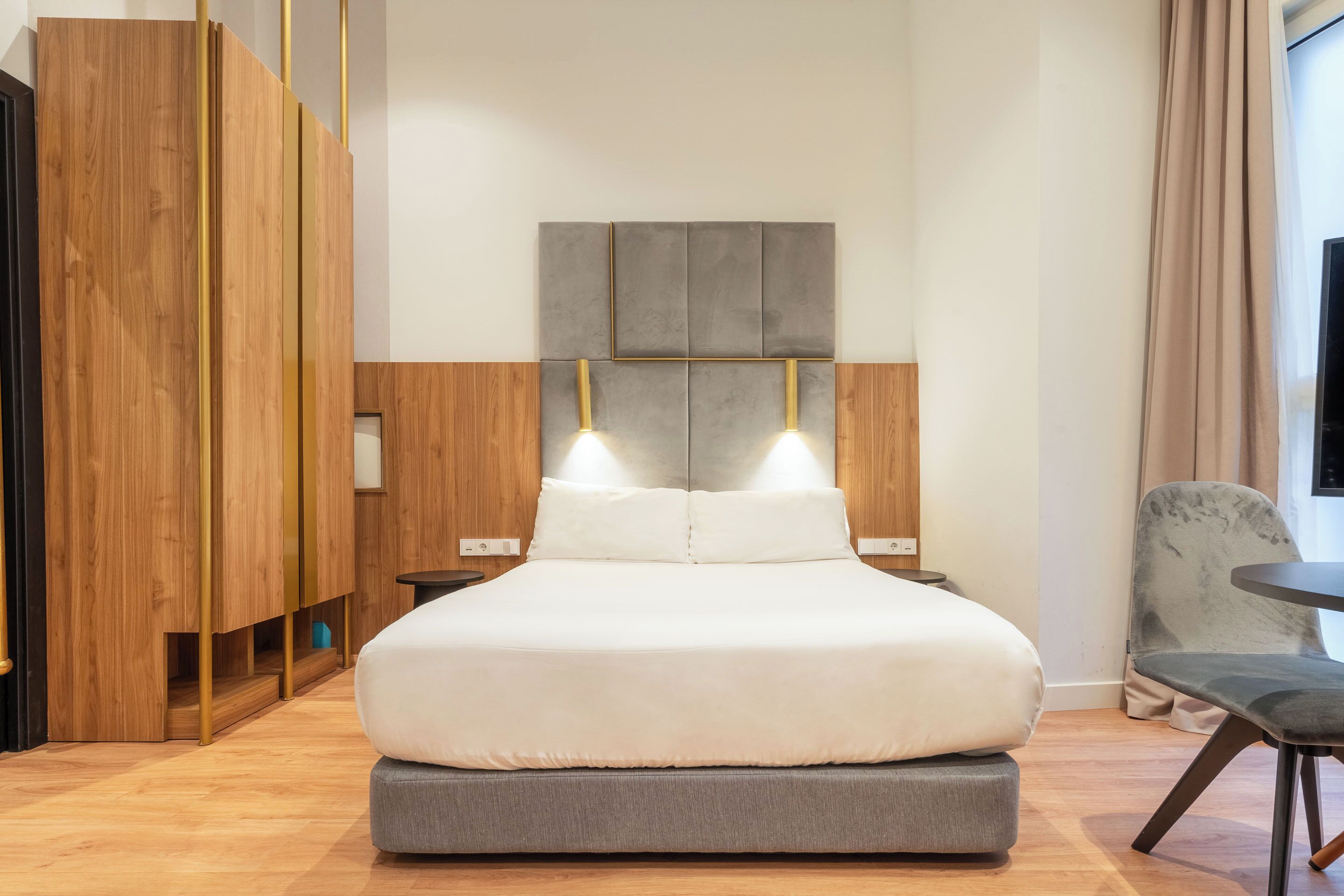 Una habitación de hotel moderna con una cama grande, un armario de madera, un sillón tapizado de color gris y una mesita.