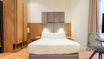 Una habitación de hotel moderna con una cama grande, un armario de madera, un sillón tapizado de color gris y una mesita.