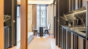 A modern hotel room with a round dining table, two chairs, a sofa, and a kitchen area with a black countertop and marble backsplash.
