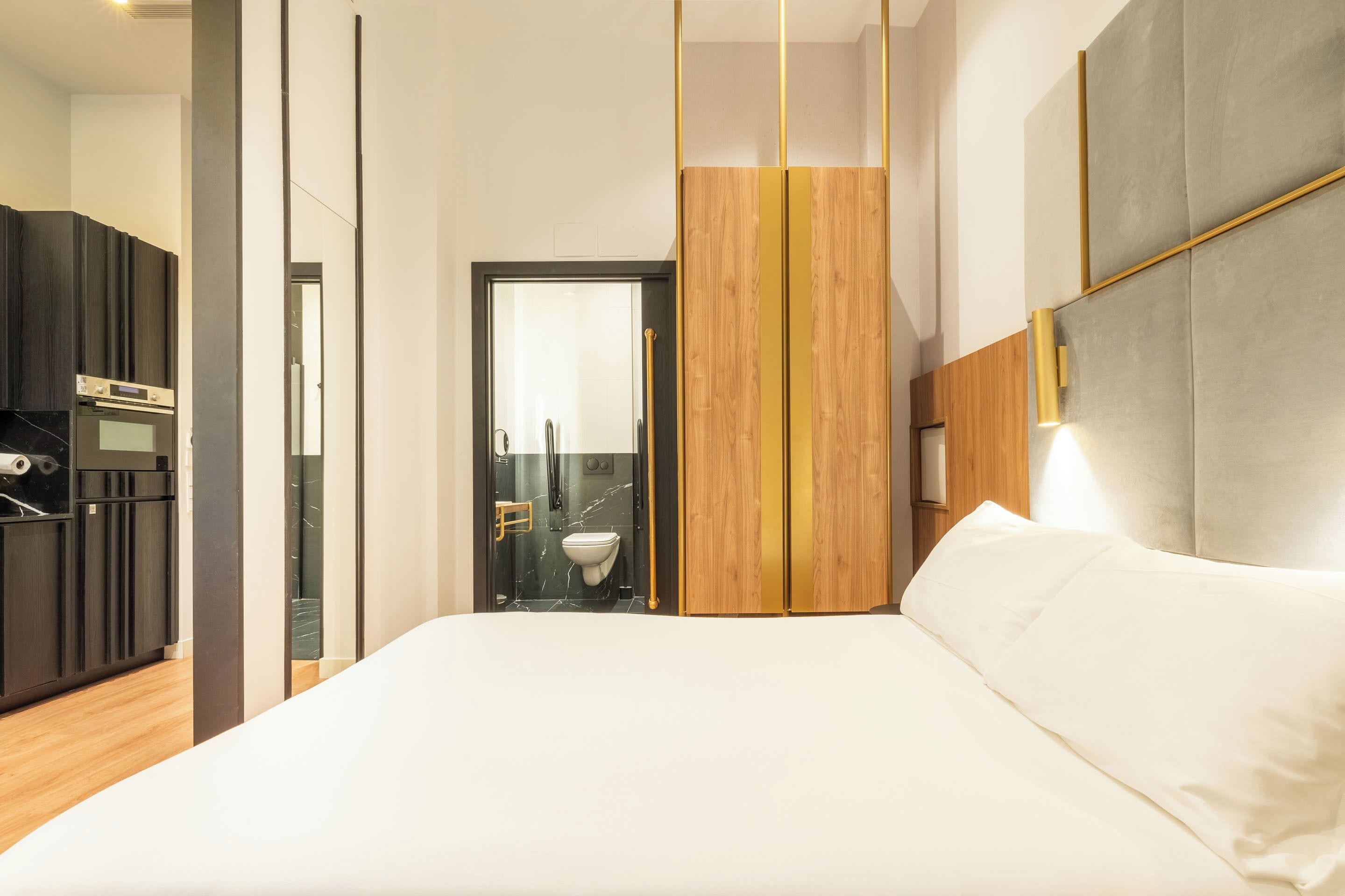 Ein modernes Hotelzimmer mit einem großen Bett, einer Holzwand, einem fest installierten Schreibtisch mit Stuhl und einem durch eine Glastür einsehbaren Badezimmer.