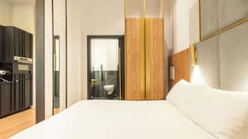 Ein modernes Hotelzimmer mit einem großen Bett, einer Holzwand, einem fest installierten Schreibtisch mit Stuhl und einem durch eine Glastür einsehbaren Badezimmer.
