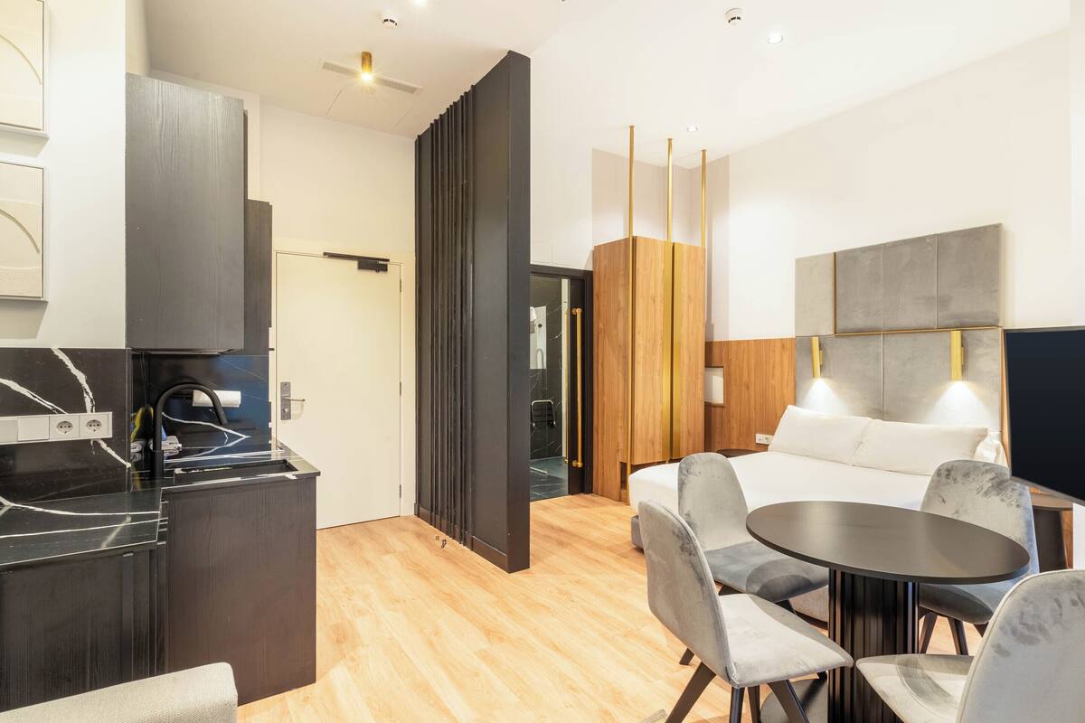 Une chambre d’hôtel moderne avec un plancher en bois, une cuisine au comptoir en marbre noir, un coin repas avec une table ronde et des chaises grises, et un grand lit avec une literie blanche.