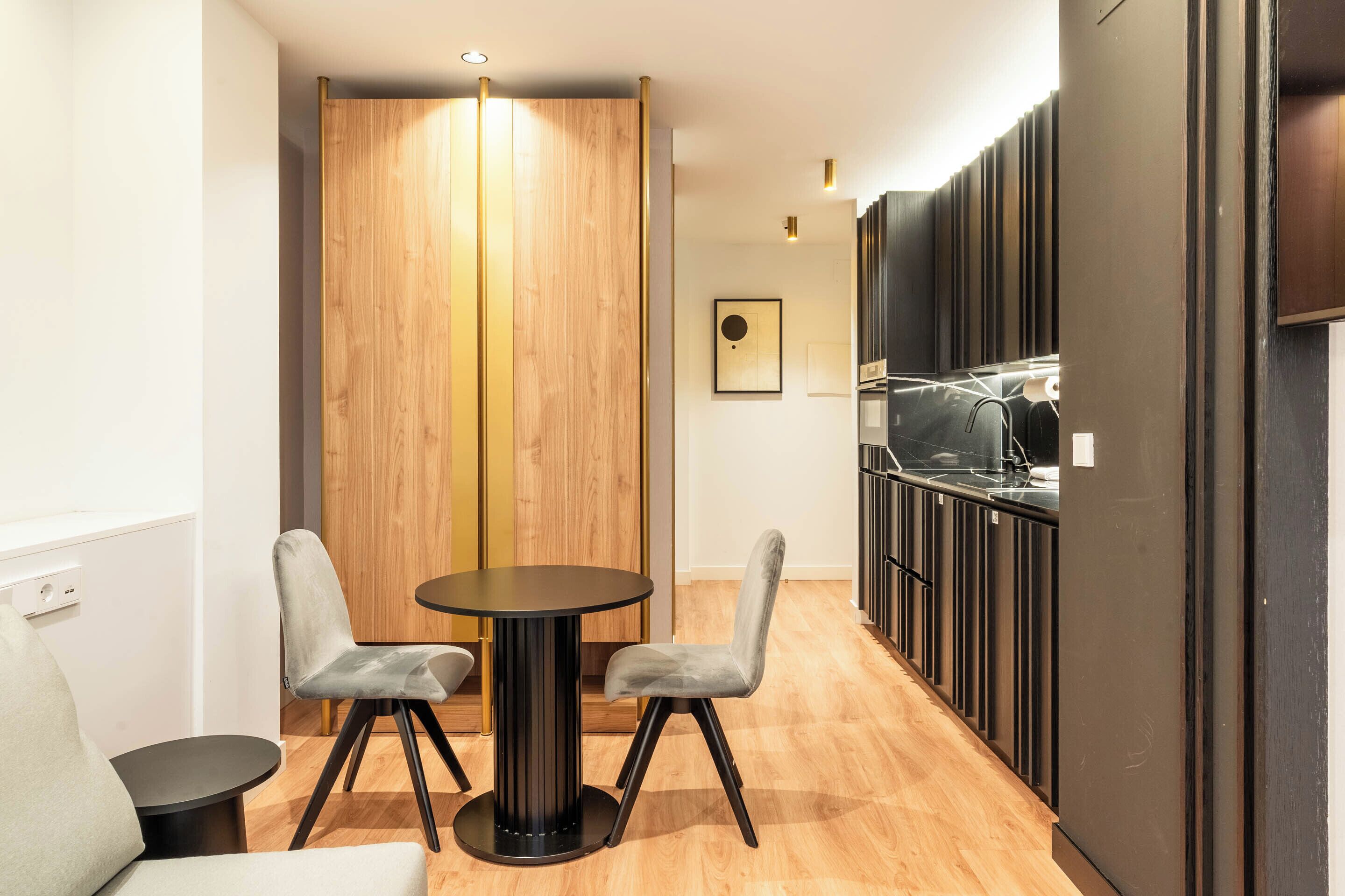 A modern dining area with a round table and two chairs, a wooden partition, and a kitchen area with black cabinets and a stainless steel sink.