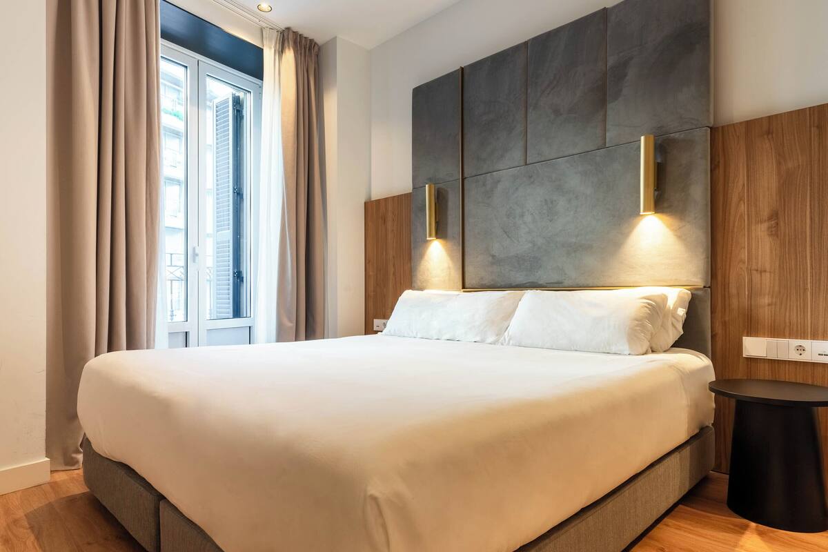Une chambre d’hôtel moderne avec un grand lit, un plancher en bois, une table de chevet et une fenêtre avec des rideaux.
