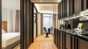 A hotel room with a bed, a small desk with a chair, a coffee machine, and a view of the dining area.