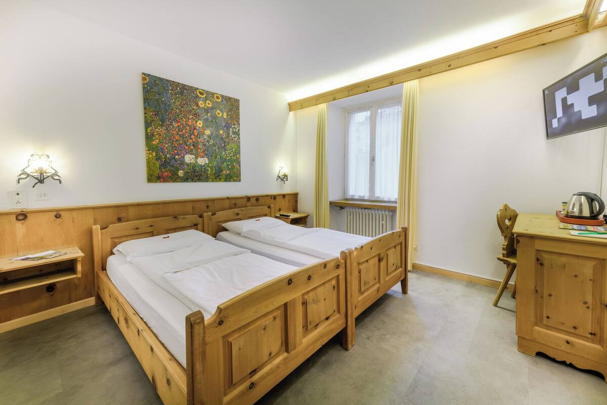 Ein hölzernes Doppelbett mit weißen Leinen, ein Nachttisch aus Holz, eine Holzkommode, ein an der Wand befestigtes Gemälde, ein Fenster mit Vorhängen und ein Spiegel.