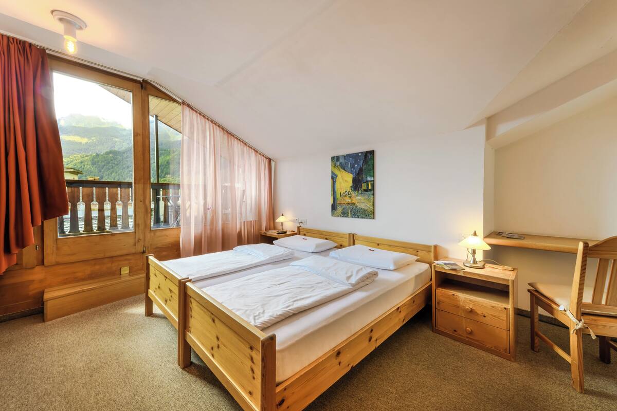 Ein hölzernes Bett mit zwei Kissen und einem Nachttisch mit Lampe. Ein Fenster mit Vorhängen, ein Bild an der Wand und ein Sessel sind ebenfalls zu sehen.