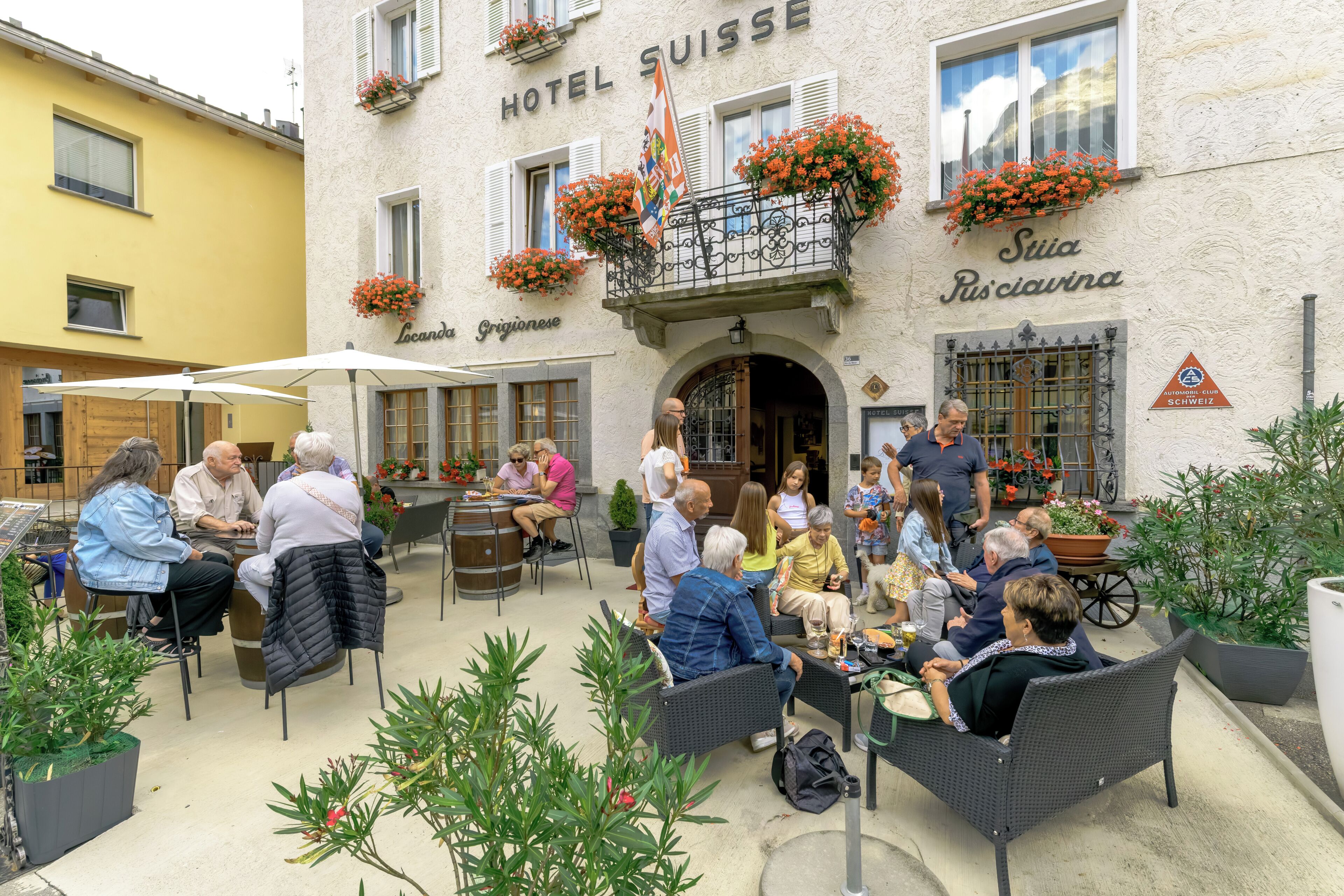 Un patio con mesas al aire libre, un edificio identificado como 'Hotel Suisse' y un letrero que anuncia cocina 'Suiza'.