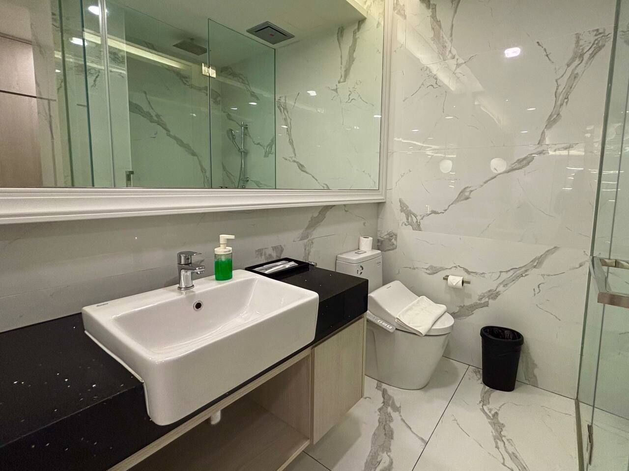 A modern bathroom with a white sink, a mirror, a toilet, and a trash bin.