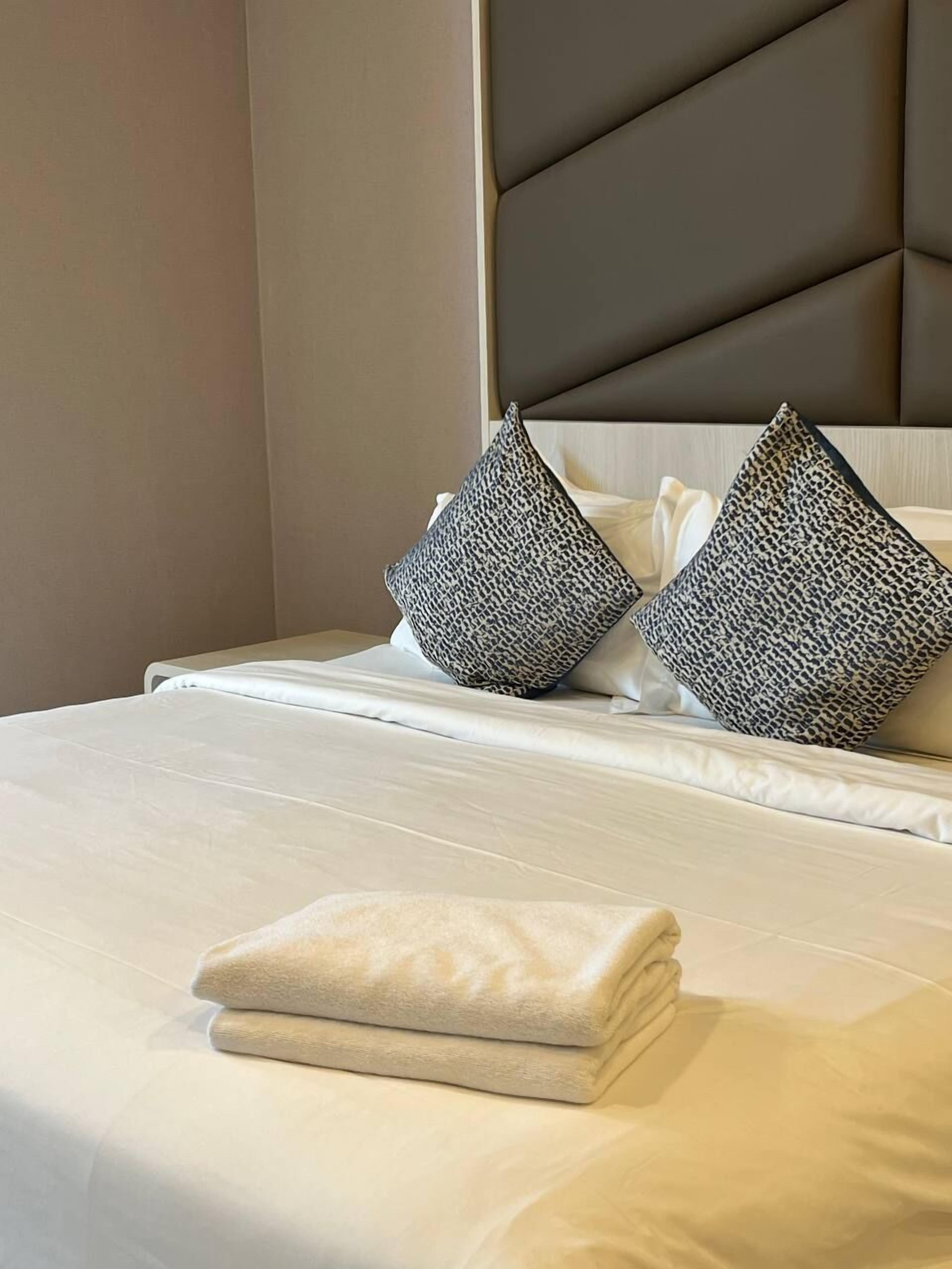 Una cama bien tendida con una toalla doblada, dos almohadas estampadas y un cabecero con diseño geométrico.