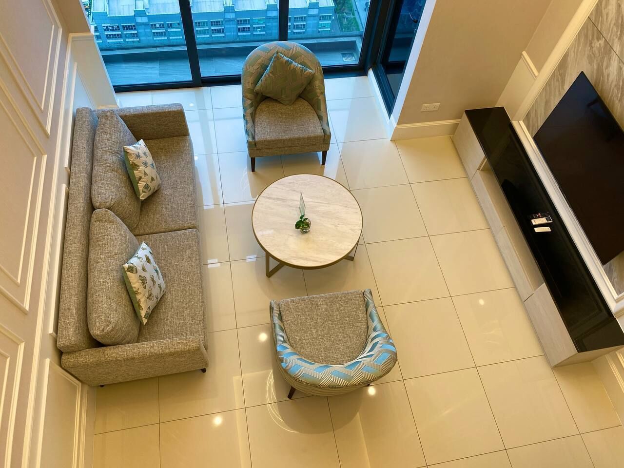 A modern living room with a sofa, armchair, and round coffee table, a fireplace, and a large window with a city view.