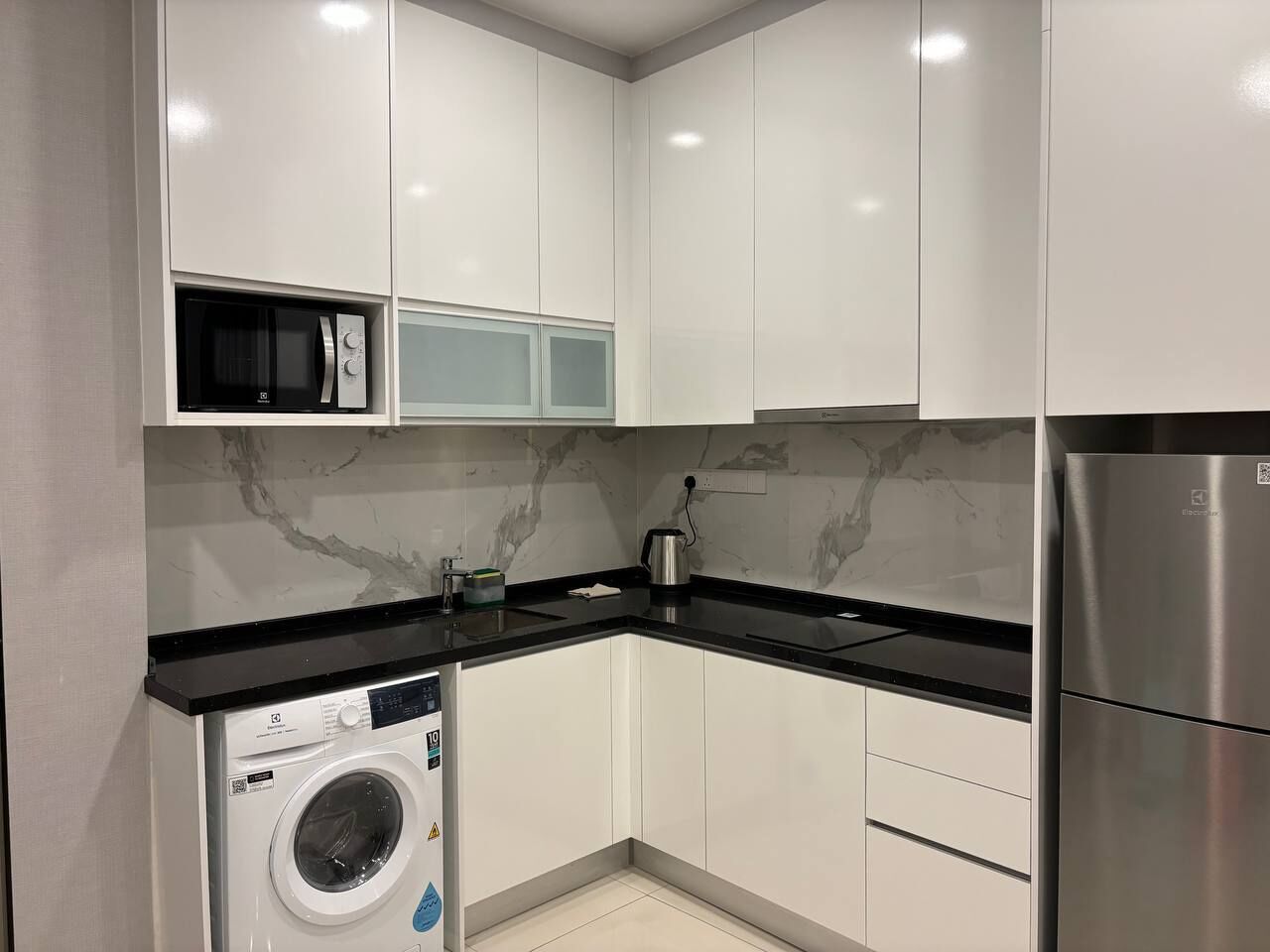 A modern kitchen with white cabinets, a stainless steel refrigerator, a washing machine, and a microwave.