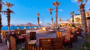 An outdoor seating area with wooden furniture and a fire pit, overlooking a body of water and mountains.