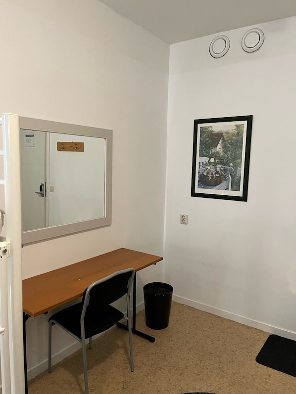 A small, clean room with a wooden desk, a black chair, a white wall with a framed picture, and a white door with a mirror. - City Hostel 46 (Örebro)