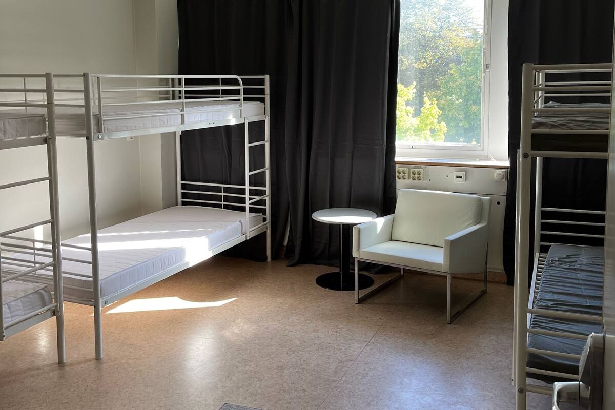 A dormitory room with bunk beds, a small round table, a chair, and a window with curtains.