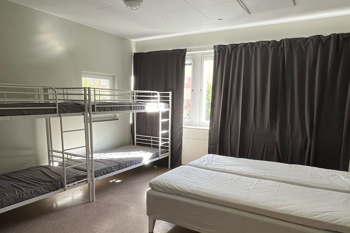 A dormitory room with bunk beds, a single bed, a window with curtains, and a ceiling with light fixtures.