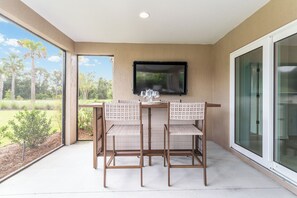 Un patio exterior con mesa y sillas, un televisor de pantalla plana montado en la pared y una puerta de vidrio que da a un jardín.