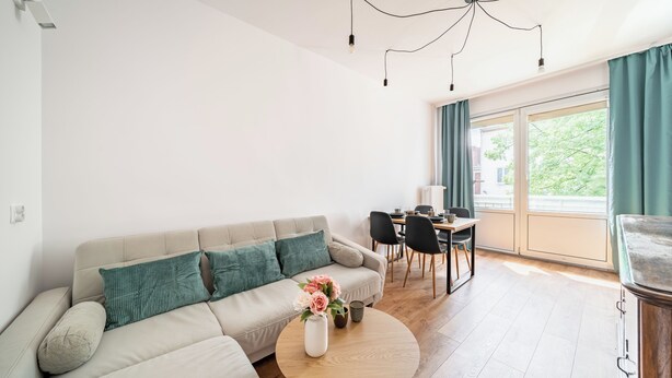 En moderne stue med sofa, spisebord med stole, en vase med blomster og udsigt til grøn vegetation udenfor.