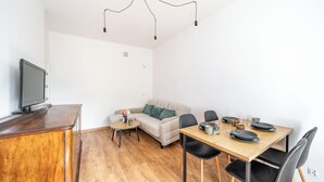 En moderne stue med trægulv, en beige sofa, et spisebord med stole, et fjernsyn på et træstel og hængende lamper.