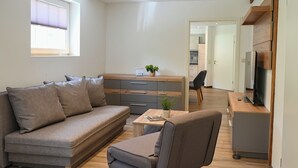 A modern living room with a sofa, a coffee table, and a TV. There is a kitchen area in the background with a window and a door leading to another room. - Romantic mill arbor "At the foot of the Staffelberg" (Bad Staffelstein)