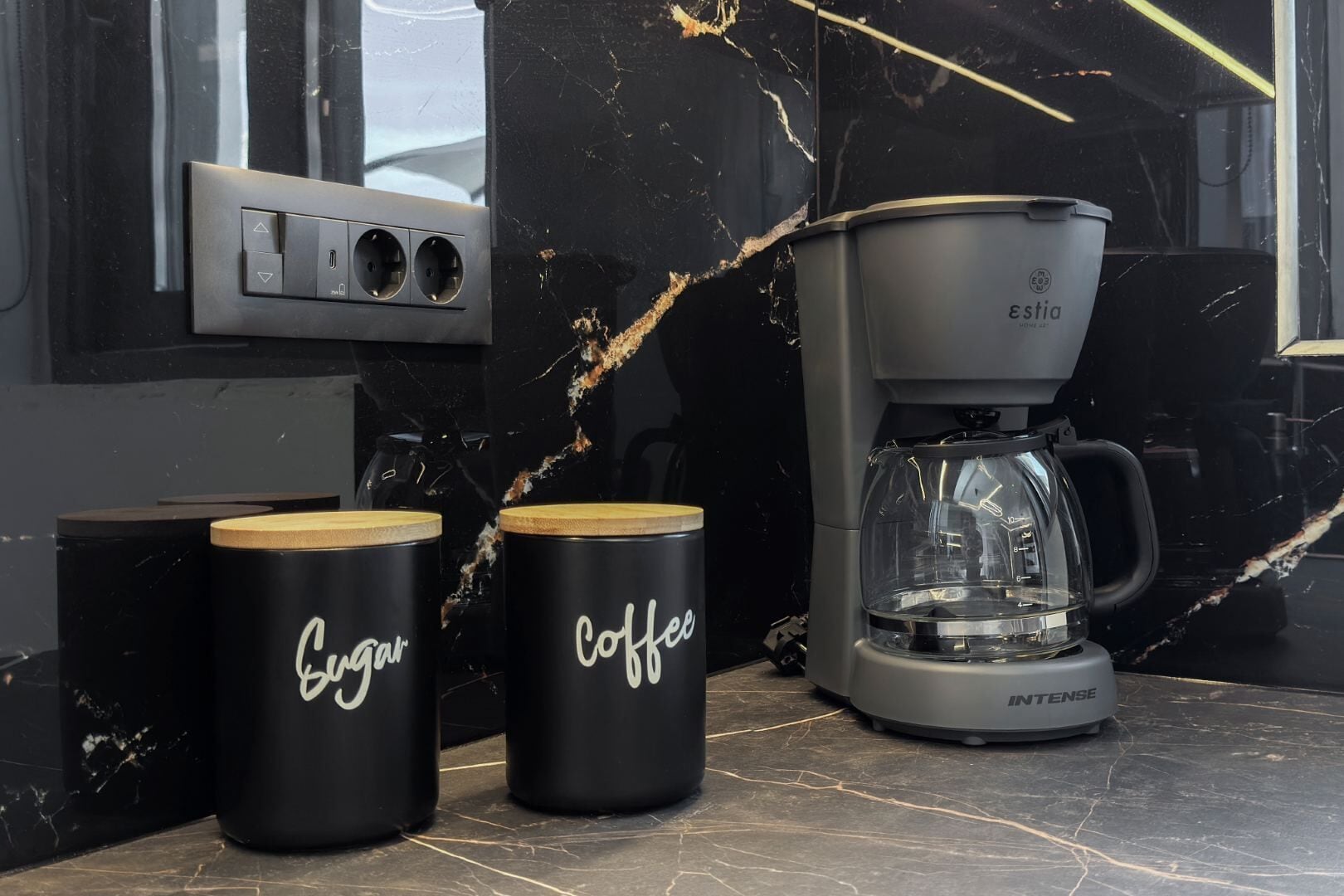 Una cocina con cafetera, dos tazas negras con la palabra 'Sugar' y 'Coffee' escritas, y un protector de salpicaduras de mármol.