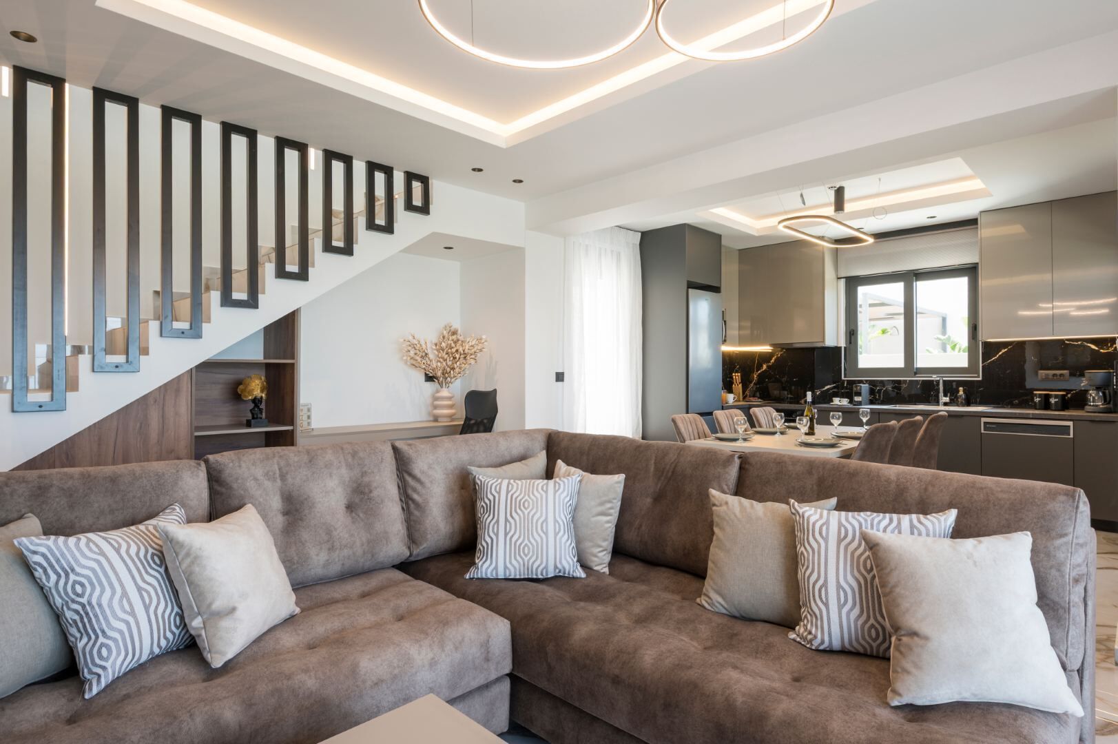 A modern living room with a sectional sofa, a staircase with framed mirrors, and a kitchen area with a dining table.