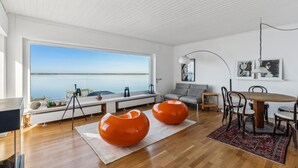 A modern living room with a large window overlooking a calm body of water, a dining area with a wooden table and chairs, and a gray sofa. - Nice home in Wunstorf/Steinhuder (Wunstorf/Steinhuder)