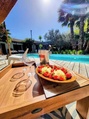 A wooden table with a tray of strawberries and bananas, a bottle of champagne, and two glasses of wine by a poolside. - "L'OLIVETTE" HOUSE: Jacuzzi, heated pool, billiards, foosball, 7 km from Nîmes (LANGLADE)