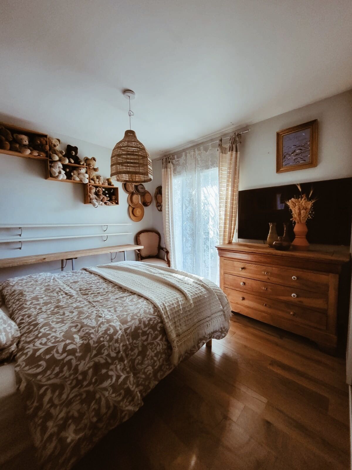 A bedroom with a bed, wooden dresser, shelves with teddy bears, a hanging light, and a TV.