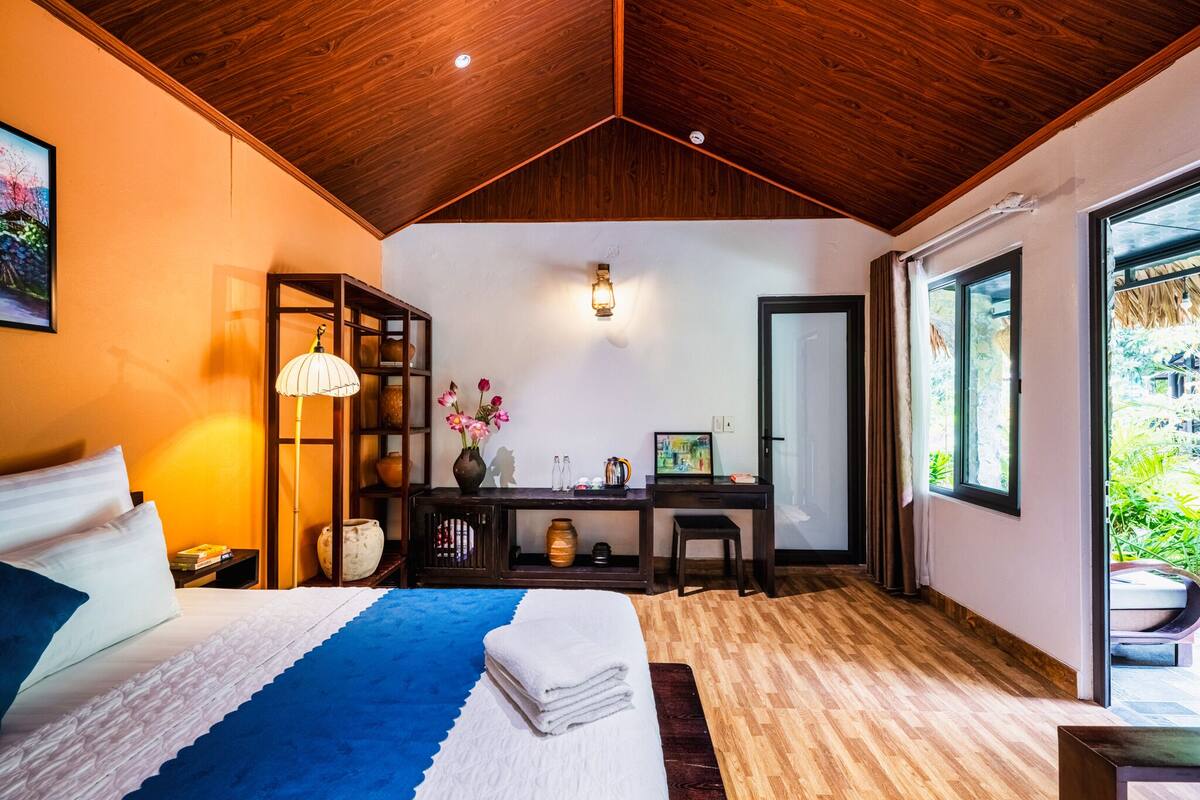A bedroom with a bed, bedside table, lamp, desk, chair, and a large window.