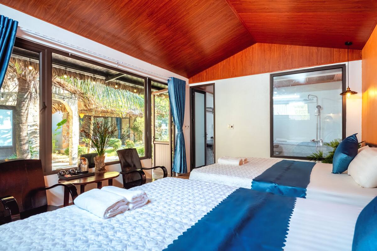 A hotel room with a wooden ceiling, large windows, a bed with a blue and white bedspread, a desk with a chair, and a view of a garden and a shower area.