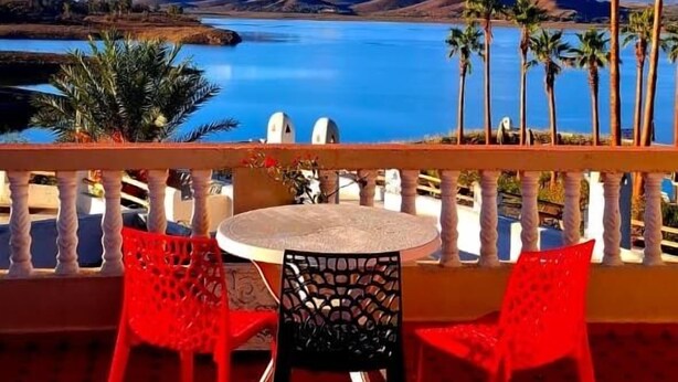 Une terrasse avec une table ronde et des chaises rouges, d’où l’on a vue sur un lac et des montagnes.
