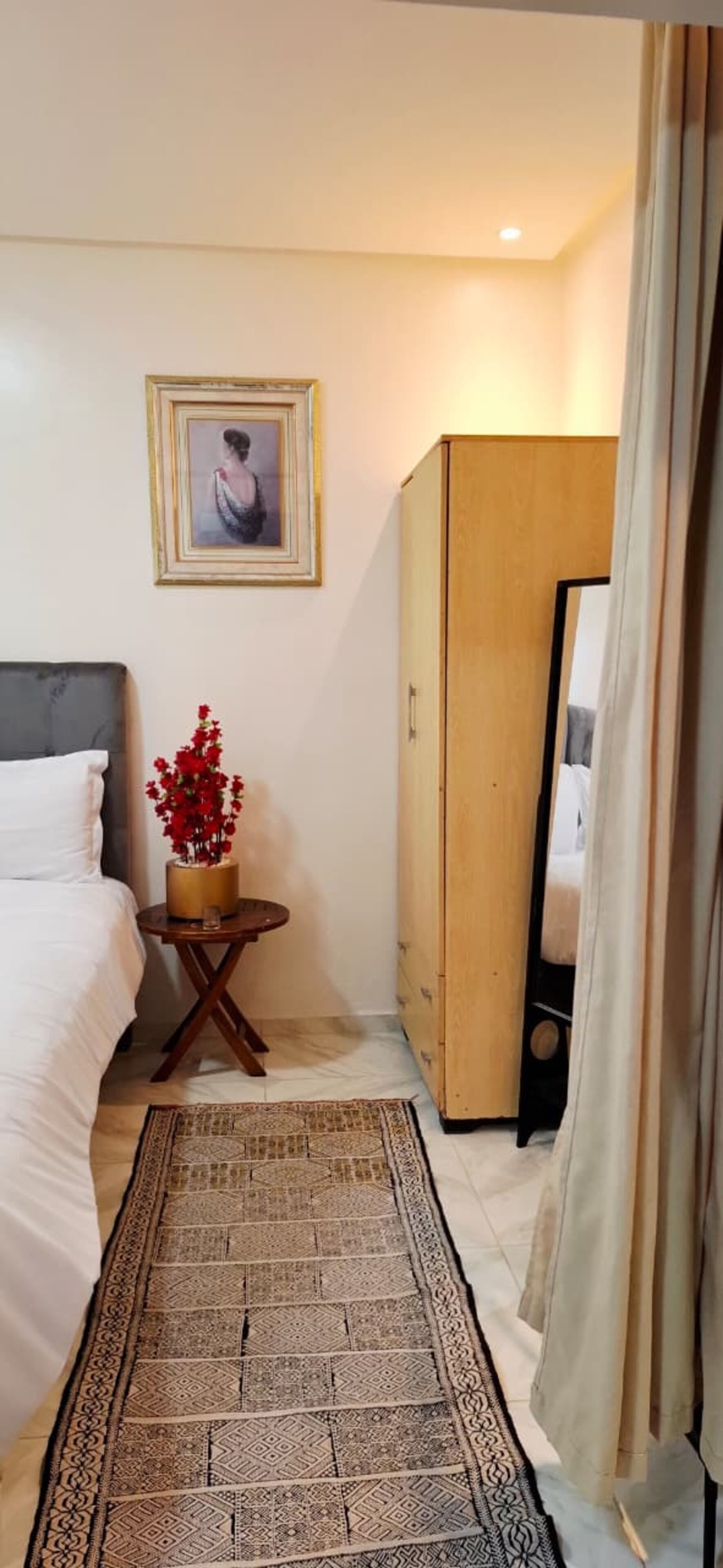 A bedroom with a bed, a wooden wardrobe, a small table with a vase of red flowers, a patterned rug, and a framed picture on the wall.