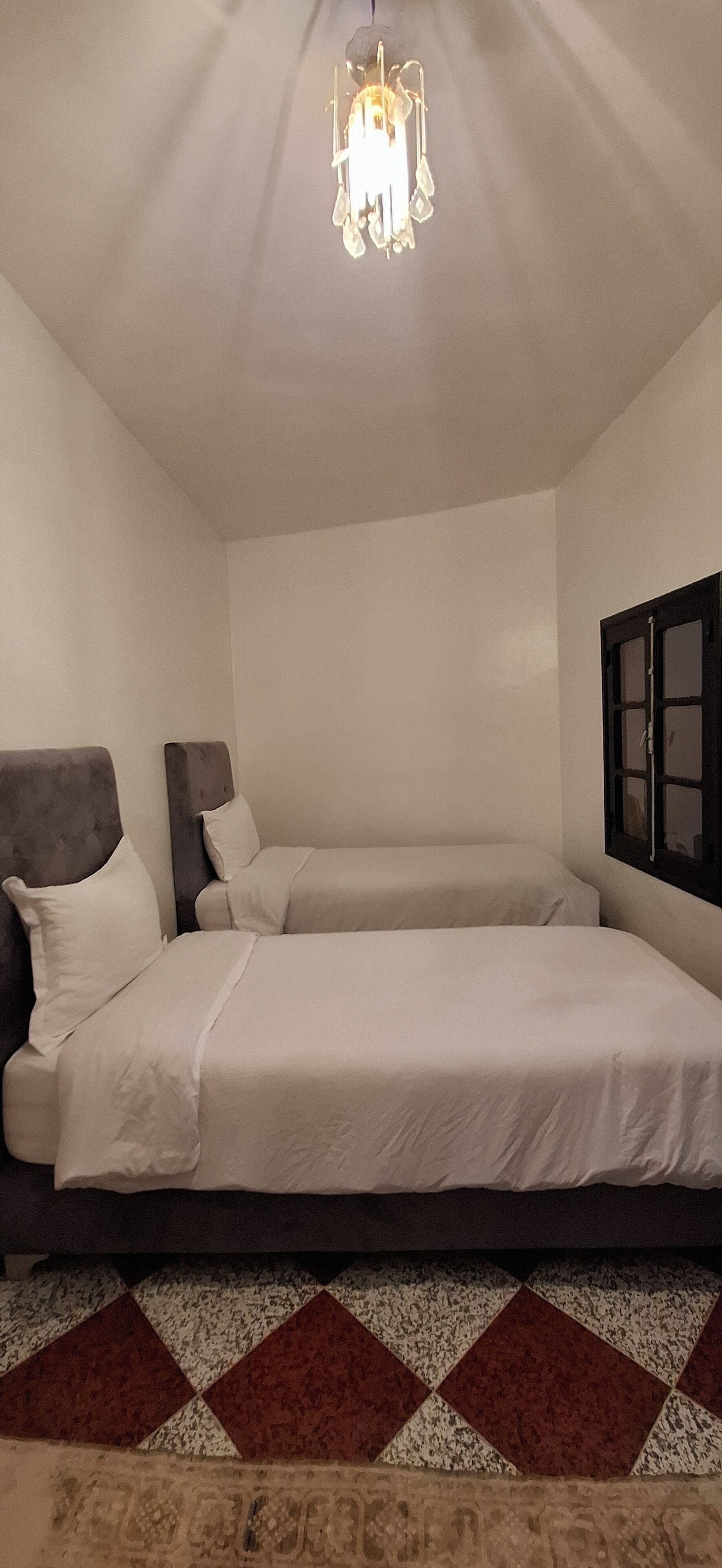 Un dormitorio con dos camas, una araña, una ventana con persianas y una alfombra estampada.