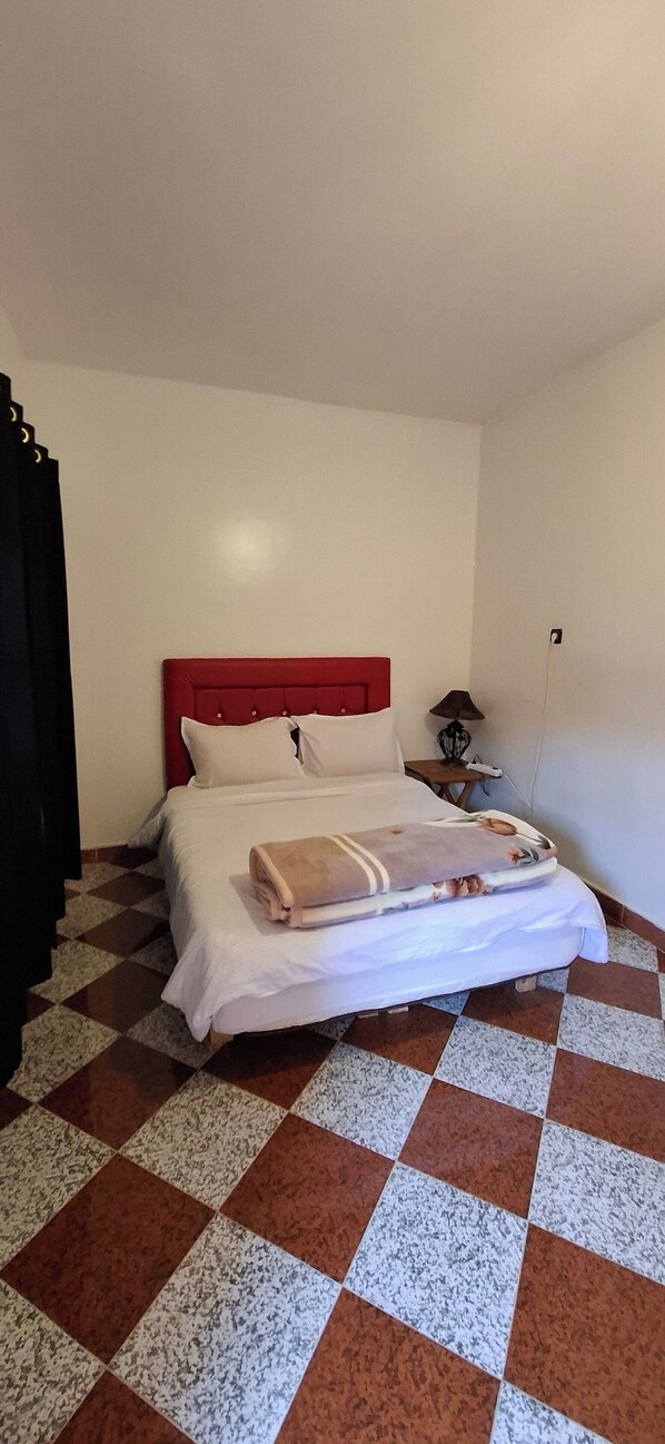 Una habitación de hotel con piso a cuadros, cabecera roja, cama con ropa blanca y mesita de noche con lámpara.