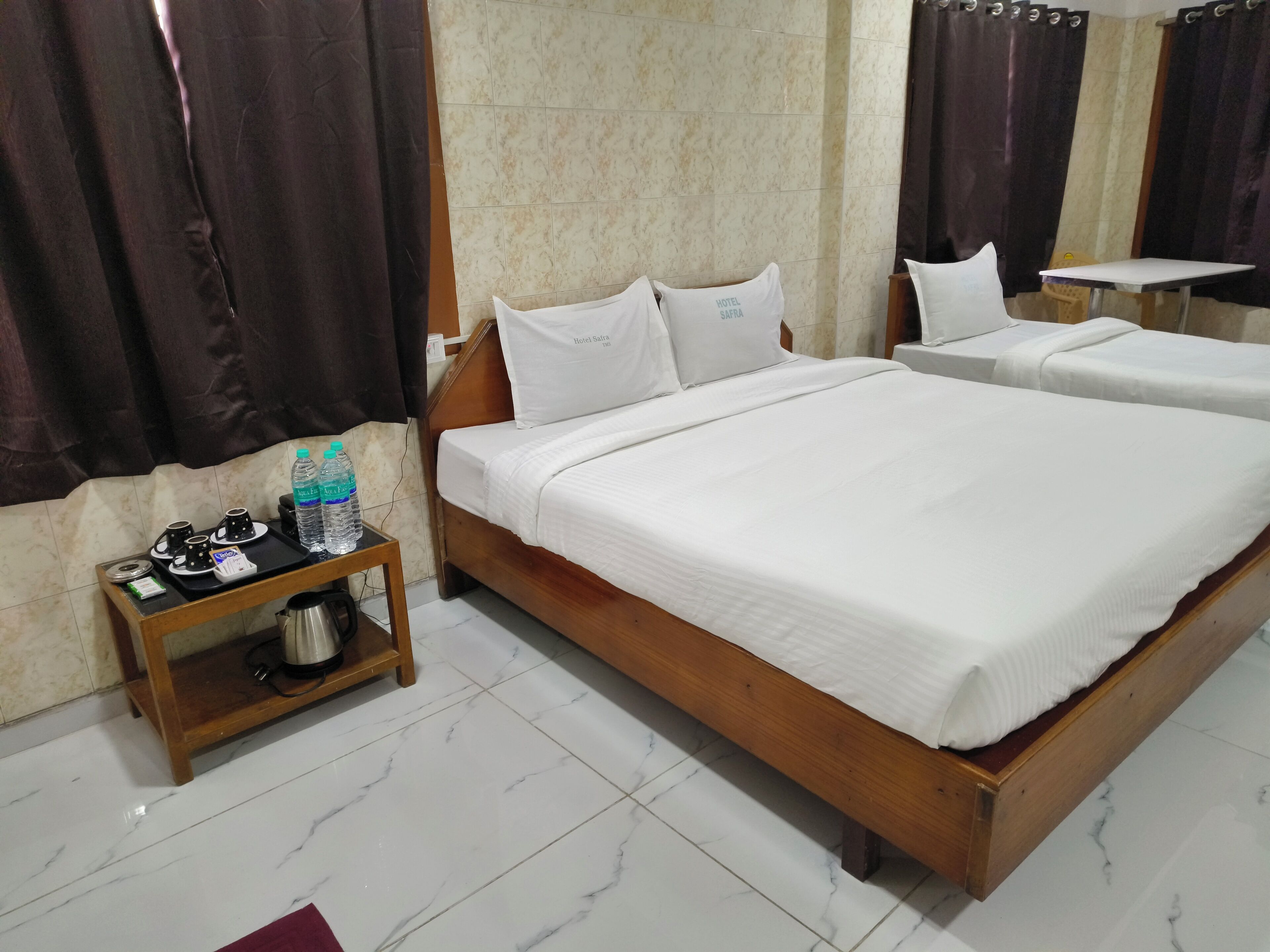 A hotel room with two beds, a small table with a kettle and a water bottle, and a tiled floor.