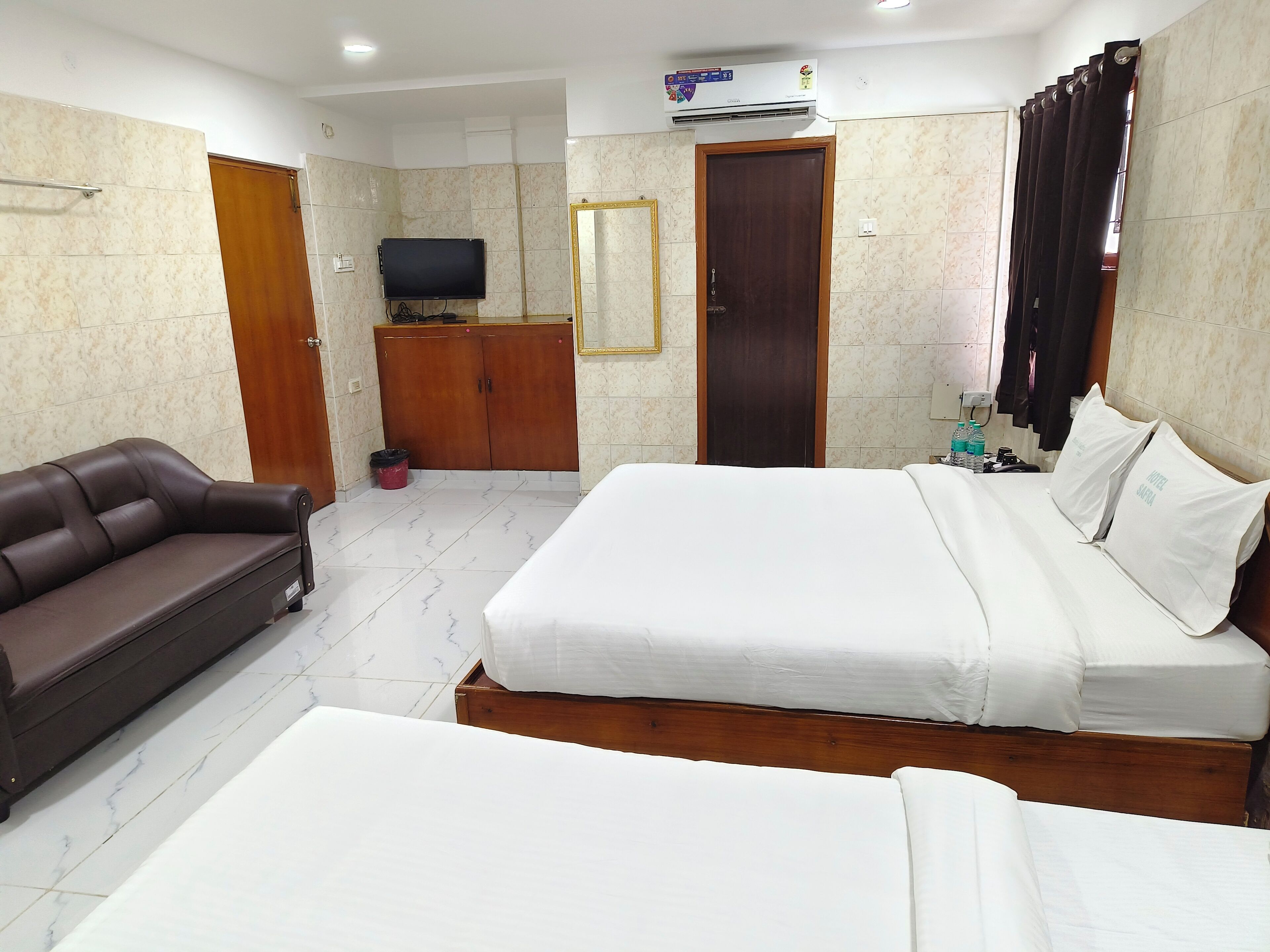 A hotel room with a large bed, a smaller bed, a sofa, a TV, a wardrobe, and an air conditioning unit.