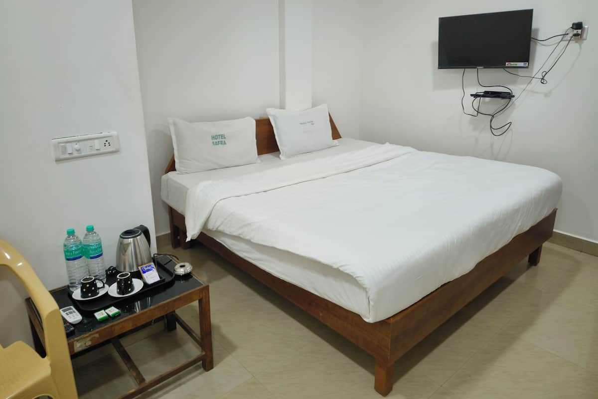 A neatly made bed with white bedding, a bedside table with a kettle, cups, and water bottles, and a TV mounted on the wall.
