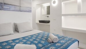 Habitación de hotel con una cama, una colcha azul estampada, almohadas blancas, una pared blanca con un cuadro, un baño con espejo y una repisa con artículos de aseo.