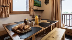Een houten eettafel met een bank, twee croissants, een fles sap en verschillende mokken met een koffielogo.